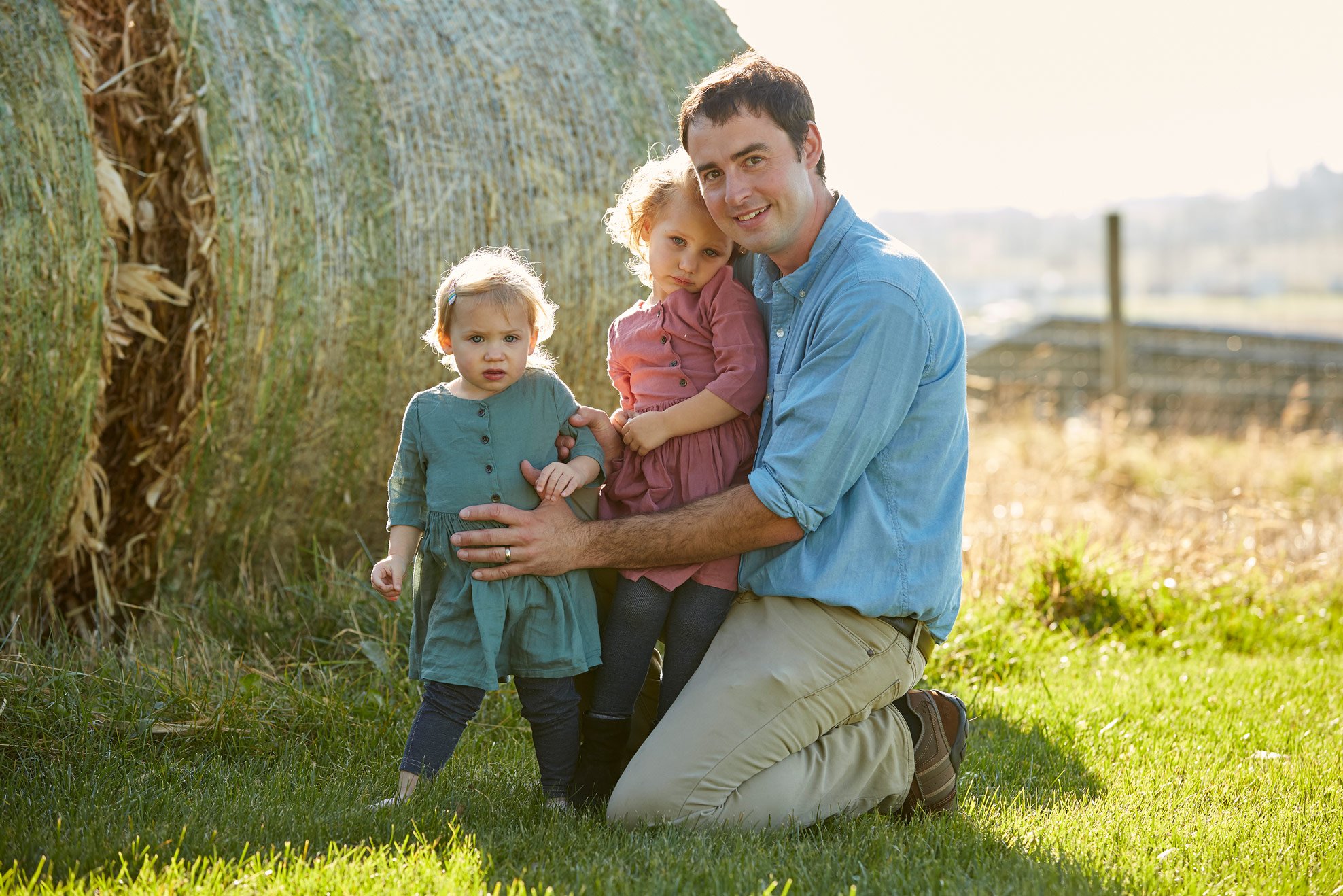 A dad with his two young daughters sitting in front of a hay bale.