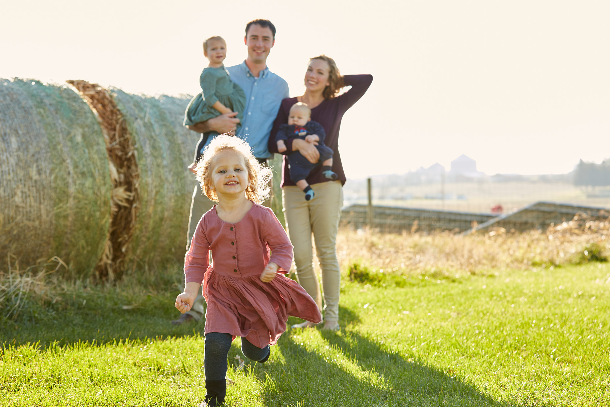 Young parents holding two children, watching their daughter run towards the camera.