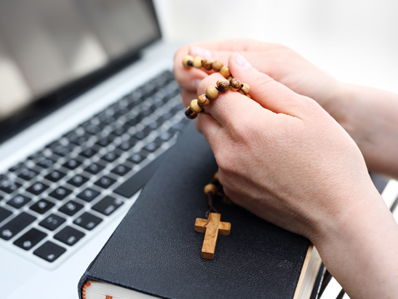 Woman holding rosary with bible and laptop below her hands